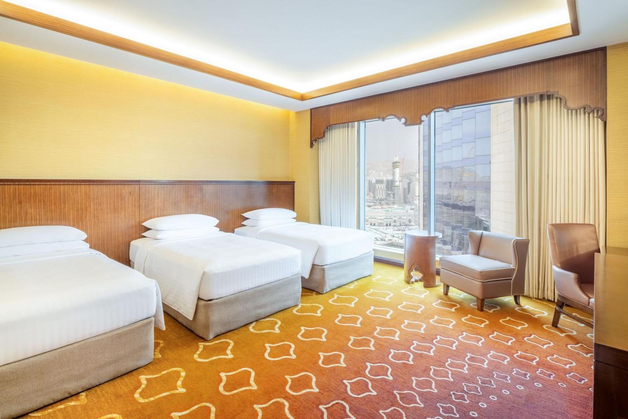 فندق جبل عمر ماريوت مكة المكرمة المظهر الخارجي الصورة The photo depicts a hotel room featuring three beds arranged in a neat manner. The walls are painted a soft yellow, and there is a decorative wooden panel behind the beds. The carpet has a unique orange design, contributing to a warm atmosphere. Larg